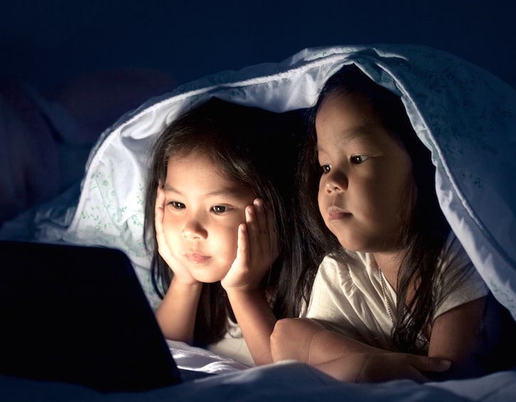 Two asian little girls using digital tablet under blanket at night