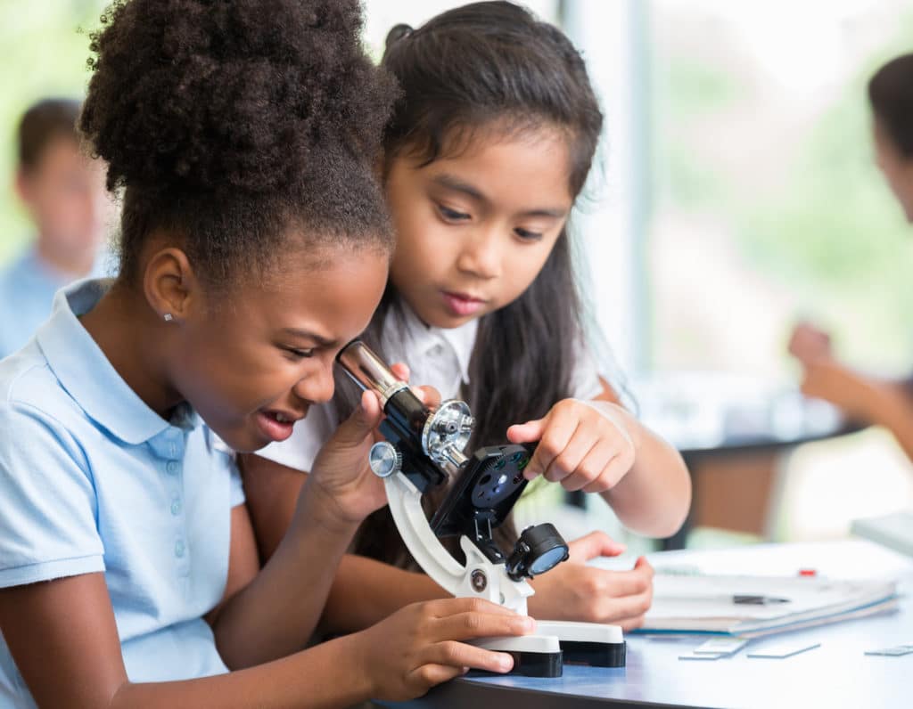 Focused African American and Asian schoolgirls help one another with their assignment in science class at a STEM elementary school. The African American girl is looking at something with a microscope.